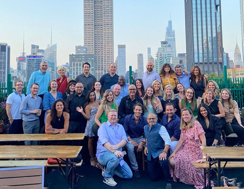 A diverse group of people pose together and smile on a rooftop with city skyscrapers in the background. Wooden tables are in the foreground, capturing the vibrant spirit of Serendipity Labs coworking on a sunny day.