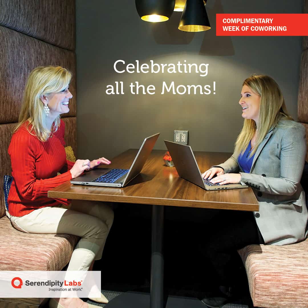 Two women sit across from each other at a booth table with laptops, smiling and talking. Text reads Celebrating all the Moms! and Complimentary Week of serendipity labs coworking. Offices and meeting rooms available. Serendipity Labs logo is in the corner.