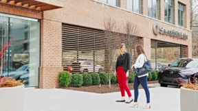 Two women walk and talk on a sidewalk outside a modern brick office building with large windows and landscaped shrubs, home to Serendipity Labs coworking offices and meeting rooms.