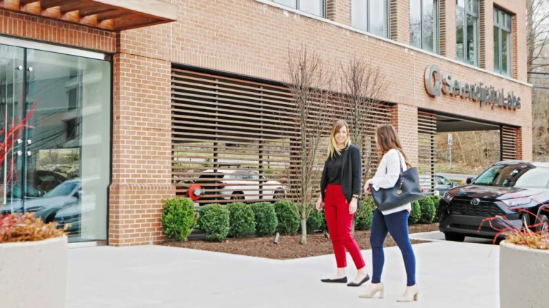 Two women walk and talk on a sidewalk outside a modern brick office building with large windows and landscaped shrubs, home to Serendipity Labs coworking offices and meeting rooms.