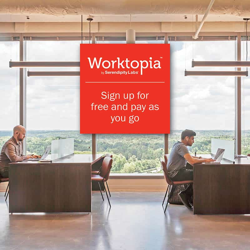 Two people work at separate desks in a modern office with large windows and scenic views, showcasing the comfort of Serendipity Labs coworking offices and meeting rooms. A red sign reads, “Worktopia. Sign up for free and pay as you go.”.