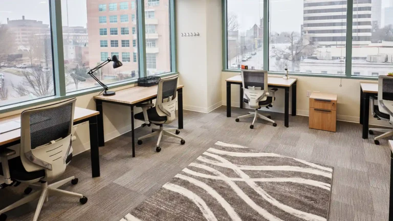A modern Serendipity Labs coworking office with large windows, three desks and chairs, a desk lamp, a small cabinet, and a gray patterned rug. The view features nearby buildings and trees on a cloudy day.