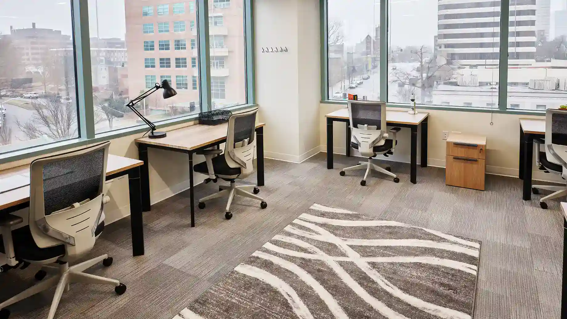 A modern Serendipity Labs coworking office with large windows, three desks and chairs, a desk lamp, a small cabinet, and a gray patterned rug. The view features nearby buildings and trees on a cloudy day.