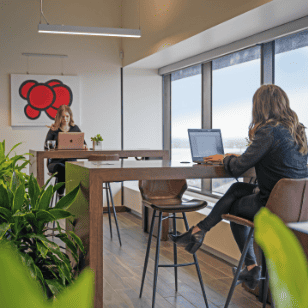 Two women work on laptops at separate wooden tables in a modern Serendipity Labs coworking space, surrounded by large windows, green plants, an abstract red art piece, and stylish offices and meeting rooms nearby.