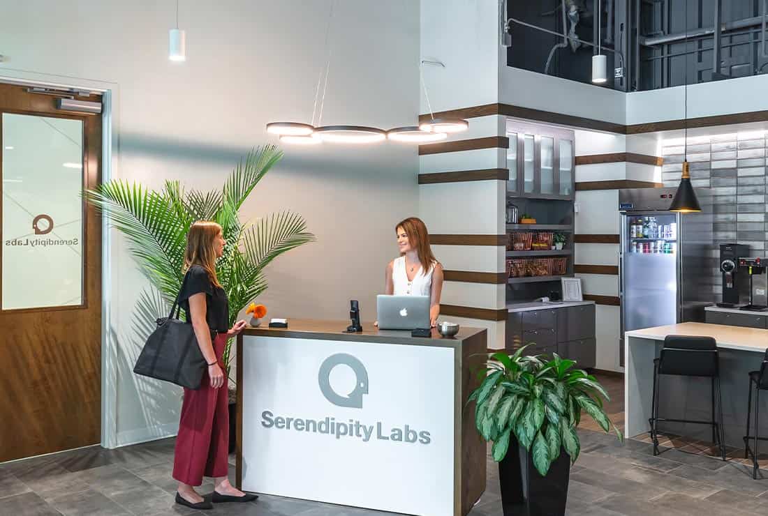A woman stands at the reception desk of Serendipity Labs coworking, speaking with a receptionist. The modern office lobby features plants, a coffee station, and stylish decor with neutral colors and wood accents.