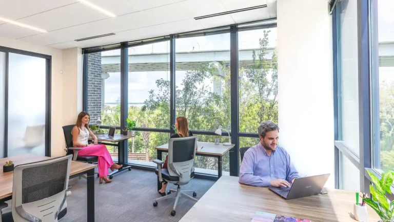 Three people work at separate desks in a bright, modern Serendipity Labs coworking space with floor-to-ceiling windows, natural light, and a view of trees. Each is focused on their laptops in the inviting offices and meeting rooms.