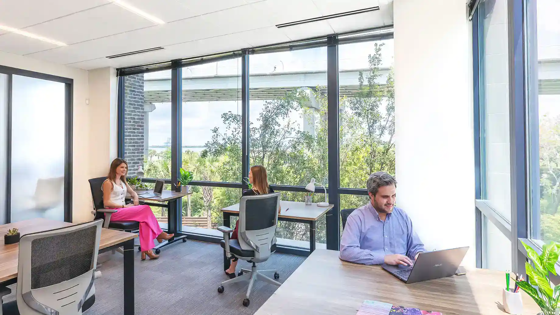Three people work at separate desks in a bright, modern Serendipity Labs coworking space with floor-to-ceiling windows, natural light, and a view of trees. Each is focused on their laptops in the inviting offices and meeting rooms.