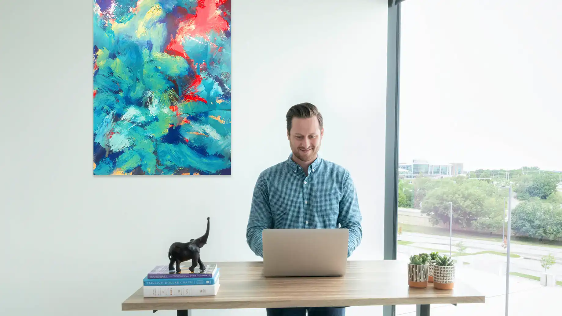A man stands at a desk using a laptop in a bright serendipity labs coworking office with large windows. Books, a small elephant statue, and plants sit on the desk, while a colorful abstract painting hangs on the wall.