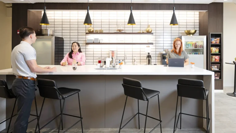 Three people are in a modern Serendipity Labs coworking office kitchen with high black chairs, a counter, pendant lights, shelves with decor, a fridge, and snacks. One is on a laptop; two converse by the counter.